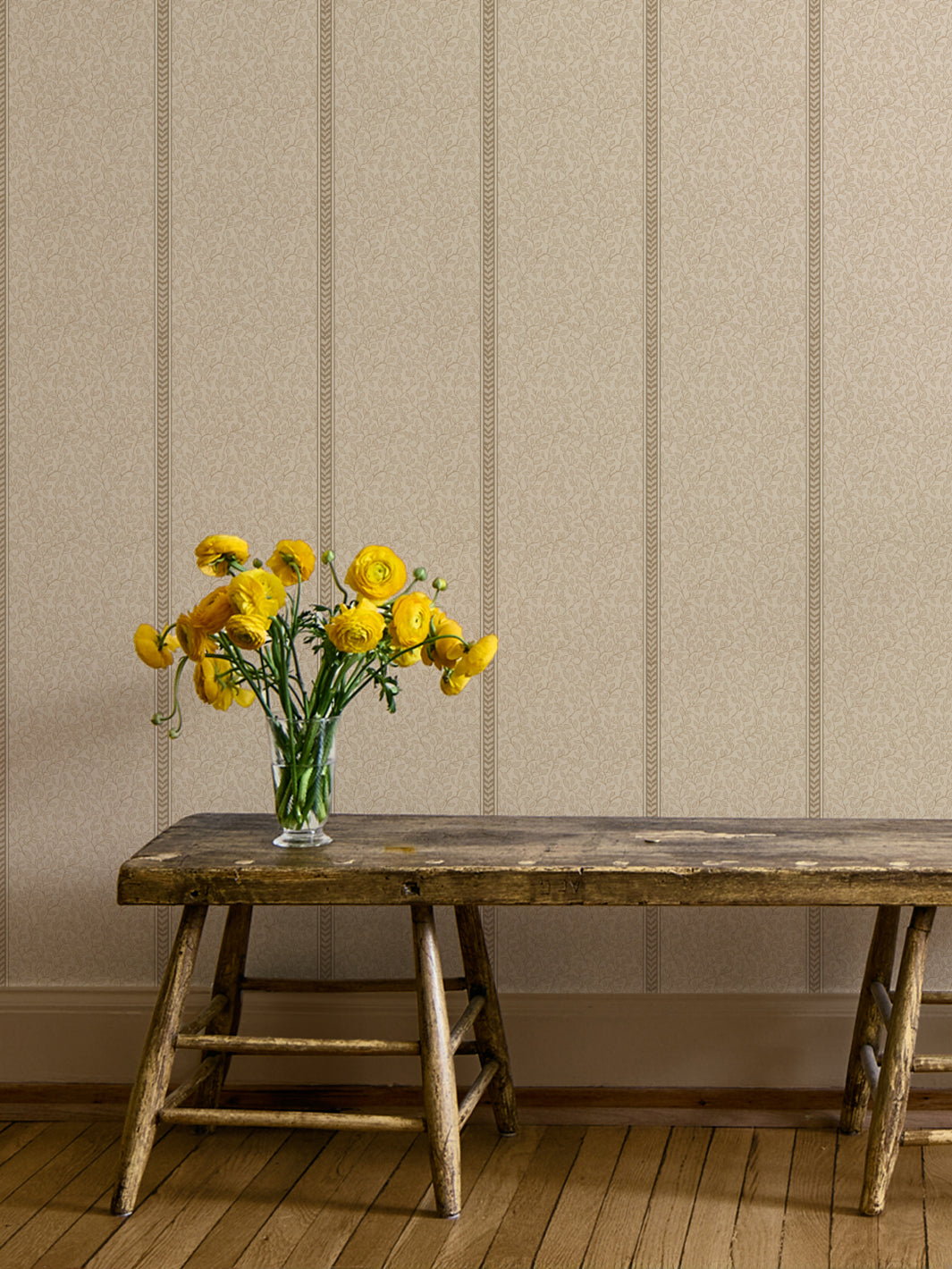 Rustic wood bench with a glass vase of yellow ranunculus against cream vine wallpaper