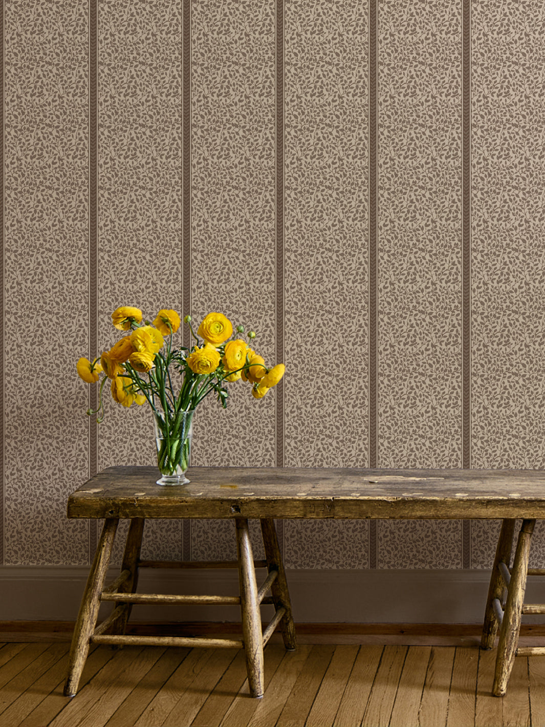 Weathered wood bench and yellow ranunculus in a glass vase against brown-on-greige vine wallpaper