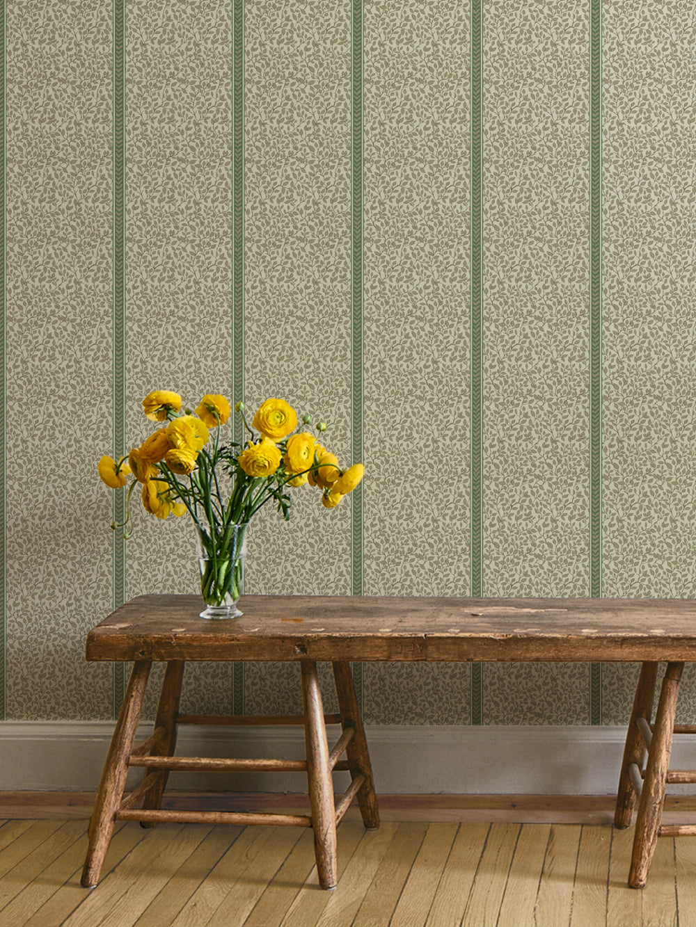 Rustic wood bench with yellow ranunculus in a glass vase against green and cream vine wallpaper