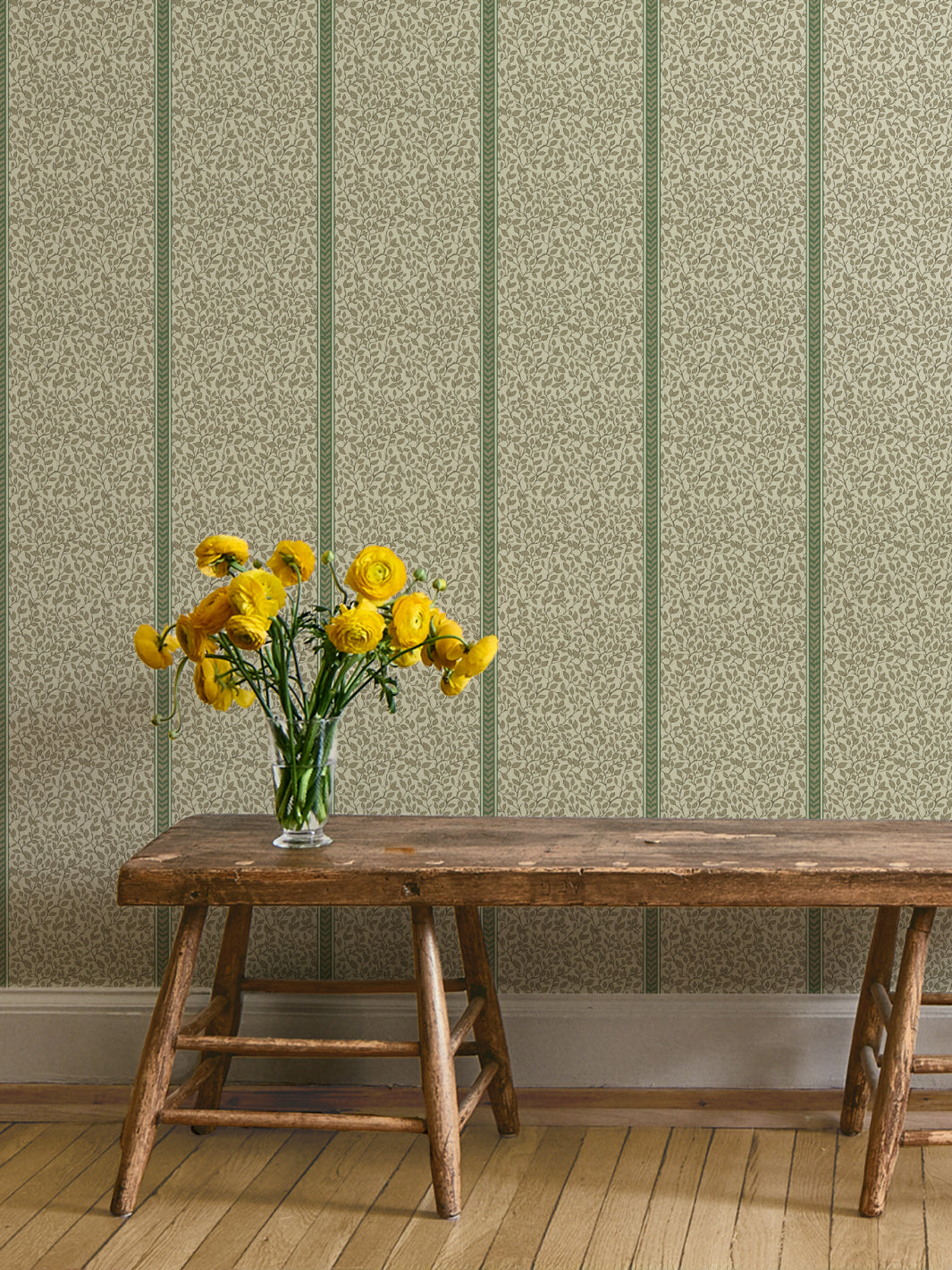 Rustic wood bench with yellow ranunculus in a glass vase against green and cream vine wallpaper