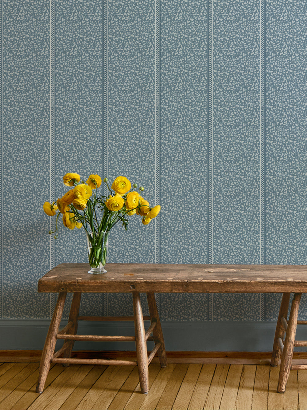 Rustic wood bench and glass vase of yellow ranunculus against blue vine wallpaper on a wood floor