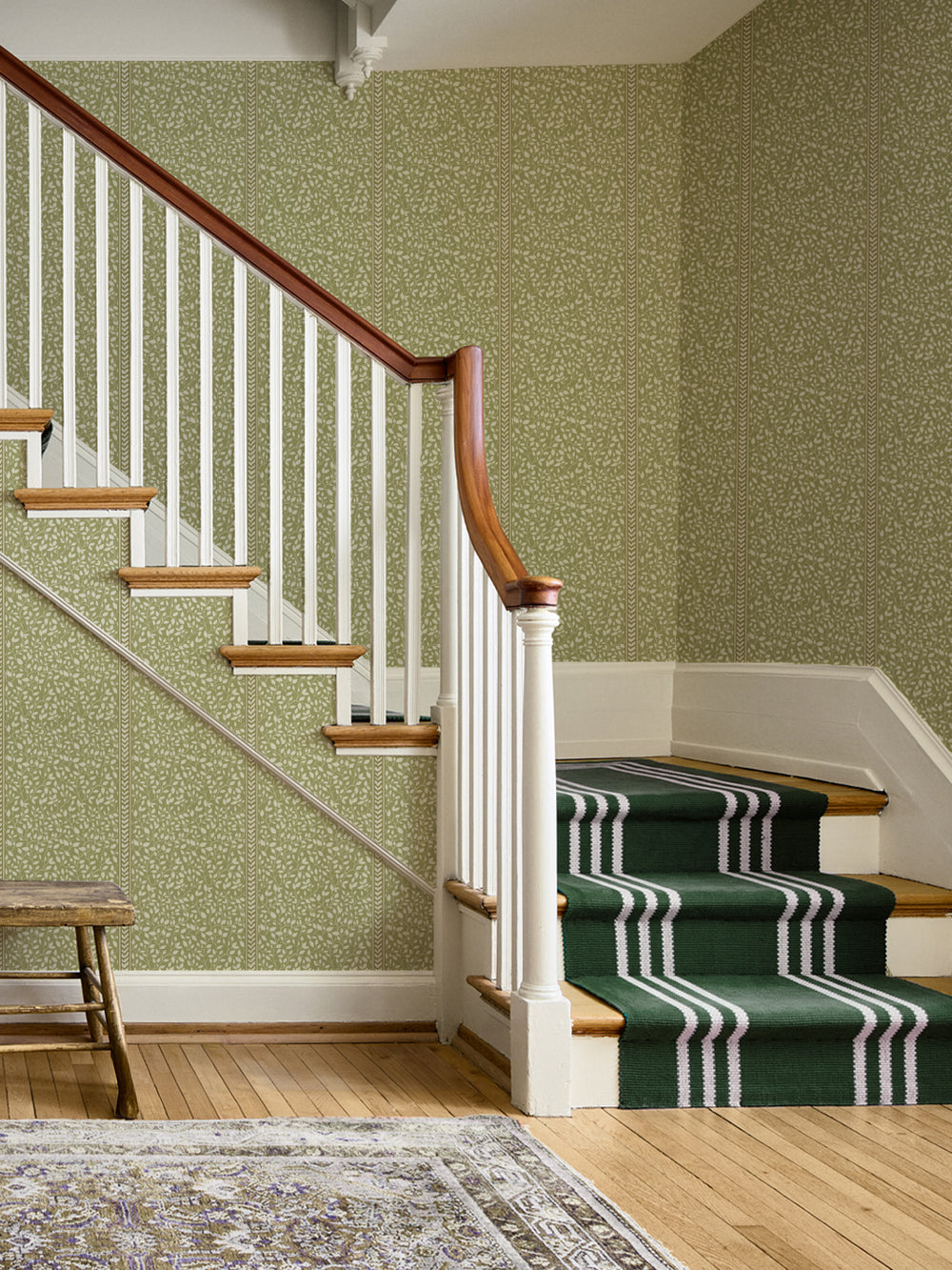 Stair hall in olive vine stripe wallpaper with white balusters, wood handrail and green stair runner