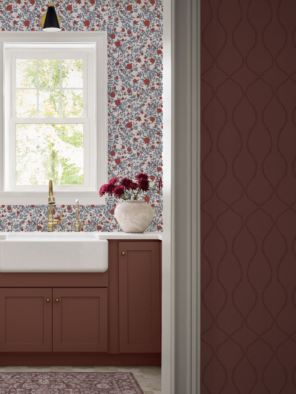 Farmhouse apron sink with a brass faucet, rust cabinets and dark red mums under a white window