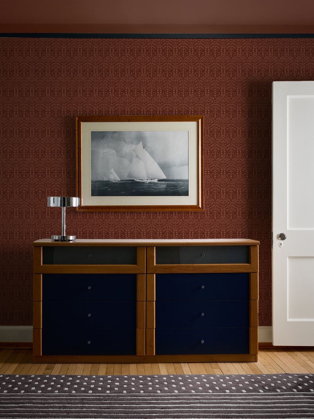 Oxblood Liliana wallpaper behind a cherry dresser with navy drawers and a framed sailboat print