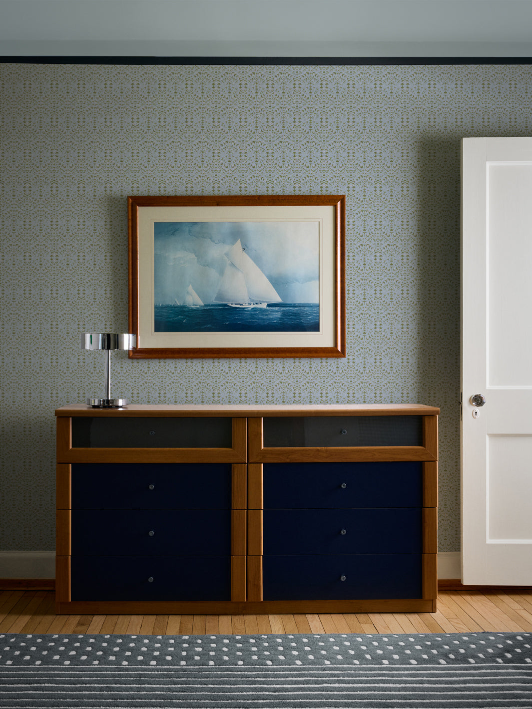 Pale blue Liliana wallpaper behind a cherry dresser with navy drawers and a framed sailboat watercolour