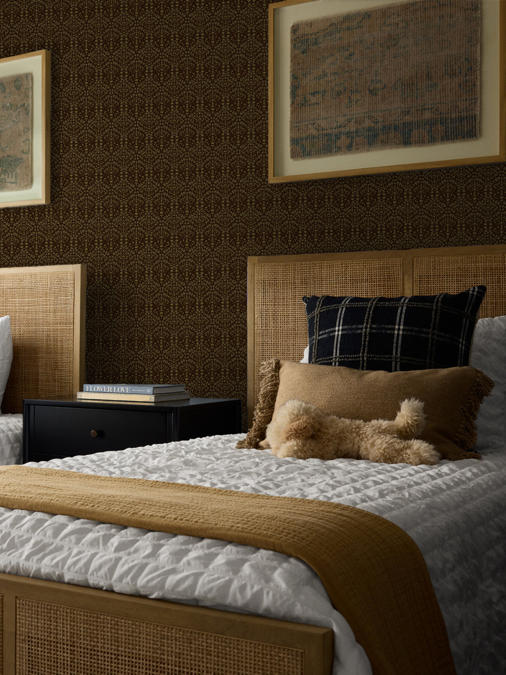Dark brown Liliana wallpaper in a bedroom with cane headboards and framed antique textiles