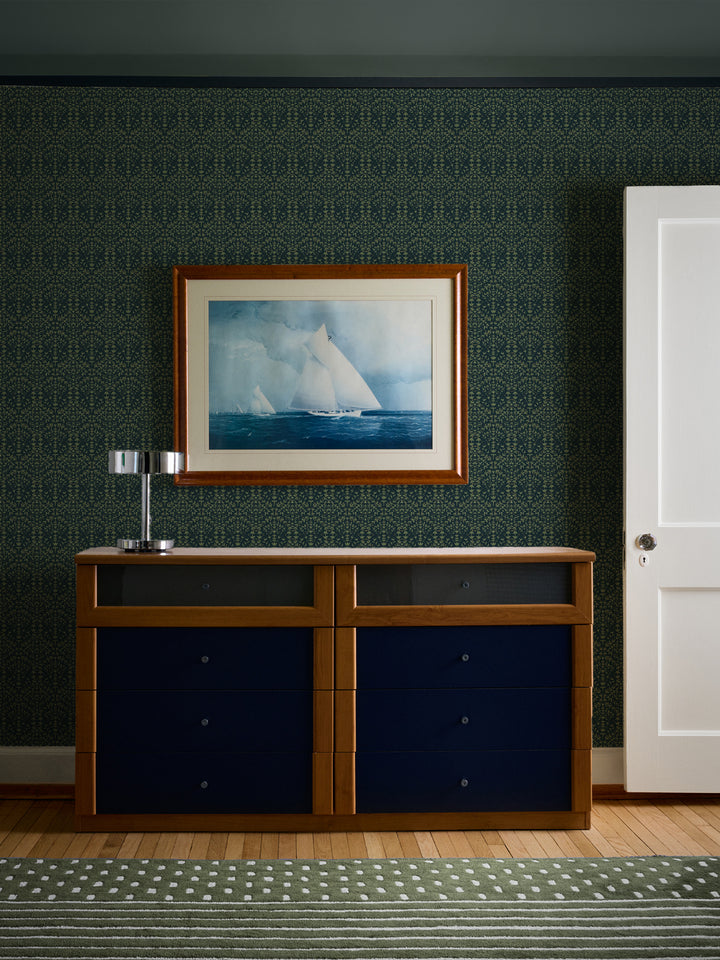 Dark green Liliana wallpaper behind a cherry dresser with navy drawers and a chrome lamp