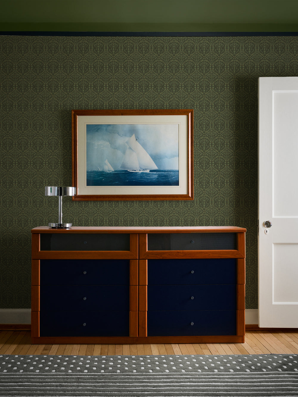 Olive-green Liliana wallpaper behind a cherry dresser with navy drawers and a framed sailboat watercolour