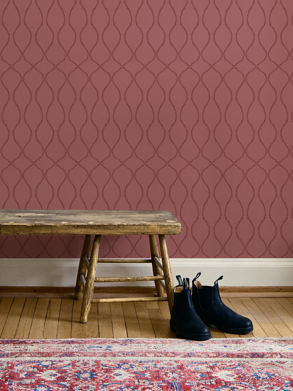 Dusty rose trellis wallpaper above a weathered wood bench with navy boots and a red oriental rug
