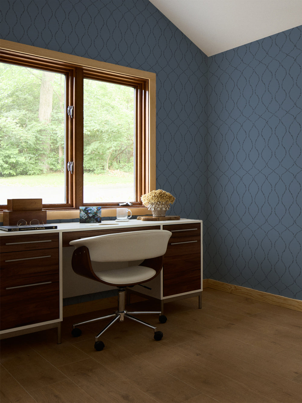 Blue trellis wallpaper in a study with a walnut desk, white swivel chair and dried hydrangeas