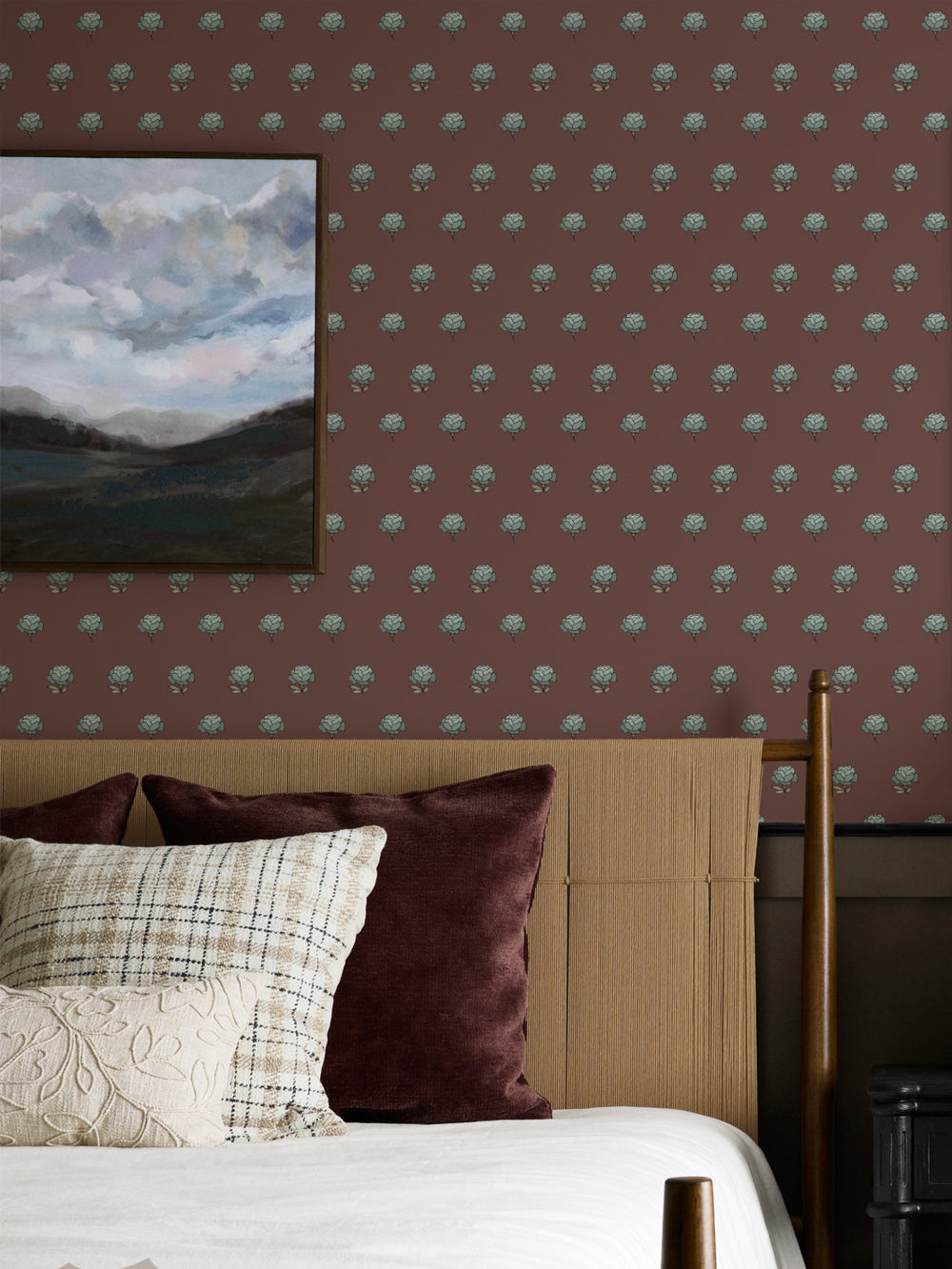Bedroom wall in maroon peony wallpaper with a cane headboard and framed cloud landscape