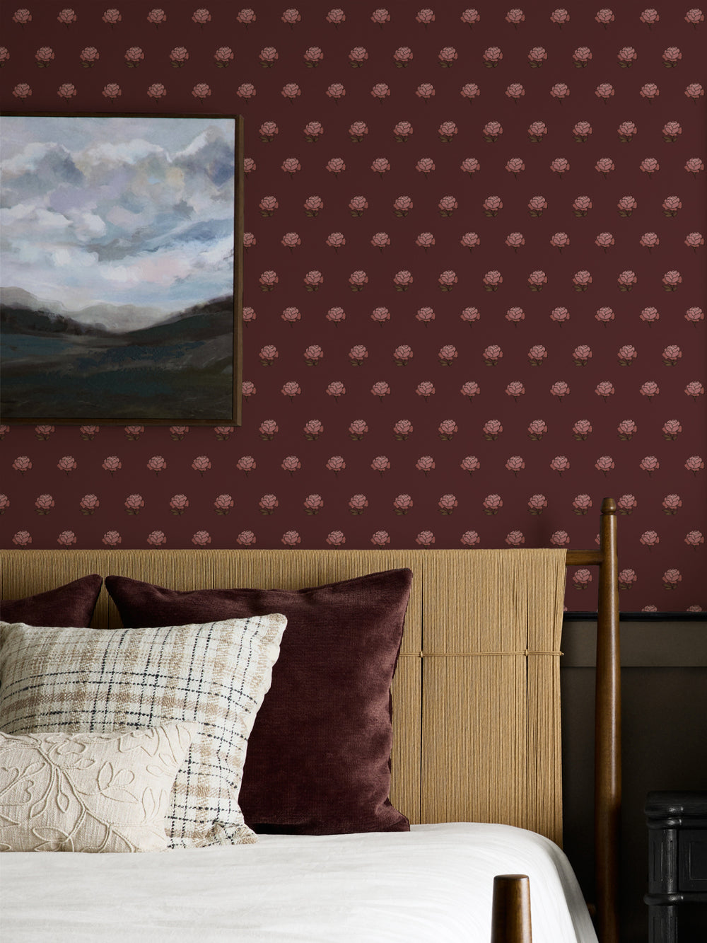 Bedroom wall in oxblood peony wallpaper with a cane headboard and framed cloud landscape