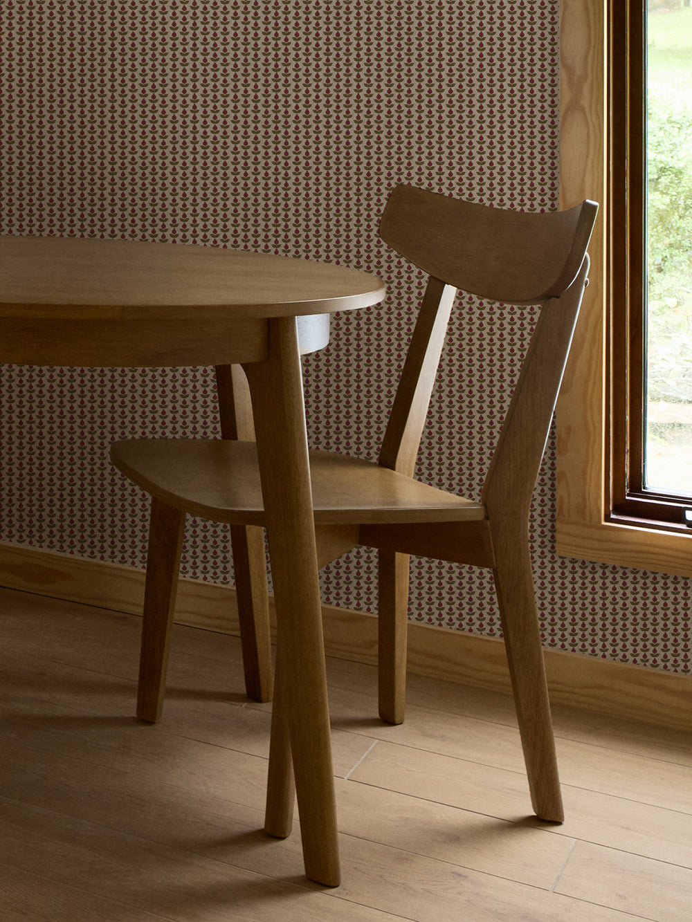 Crimson and olive Pomara Bloom wallpaper behind a round oak dining table and chair by a window