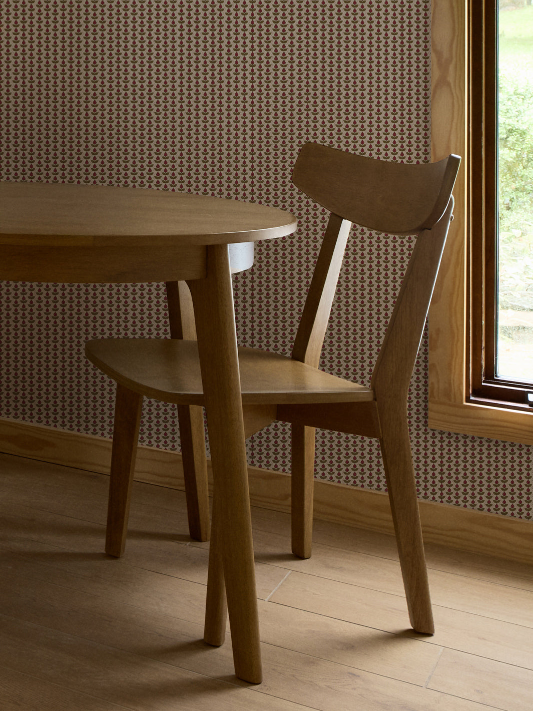Crimson and olive Pomara Bloom wallpaper behind a round oak dining table and chair by a window