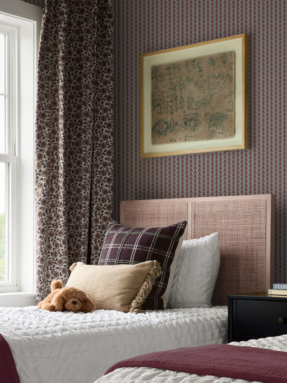Slate-blue Pomara Bloom wallpaper in a bedroom with a pink cane headboard and burgundy throw