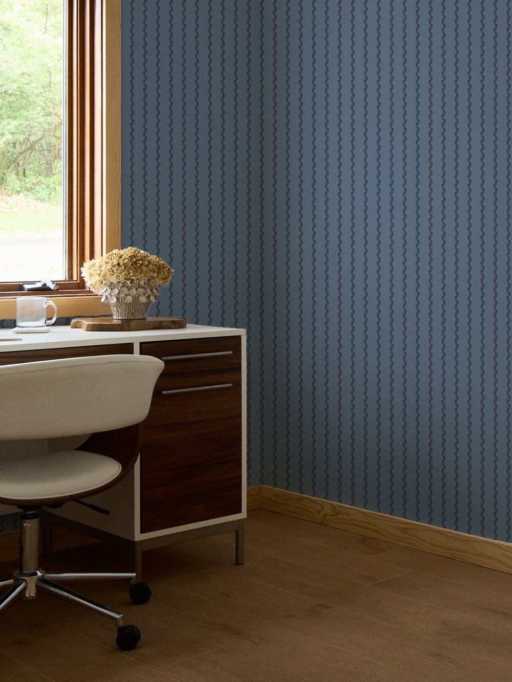 Slate blue stripe wallpaper behind a walnut desk with a white swivel chair by a window