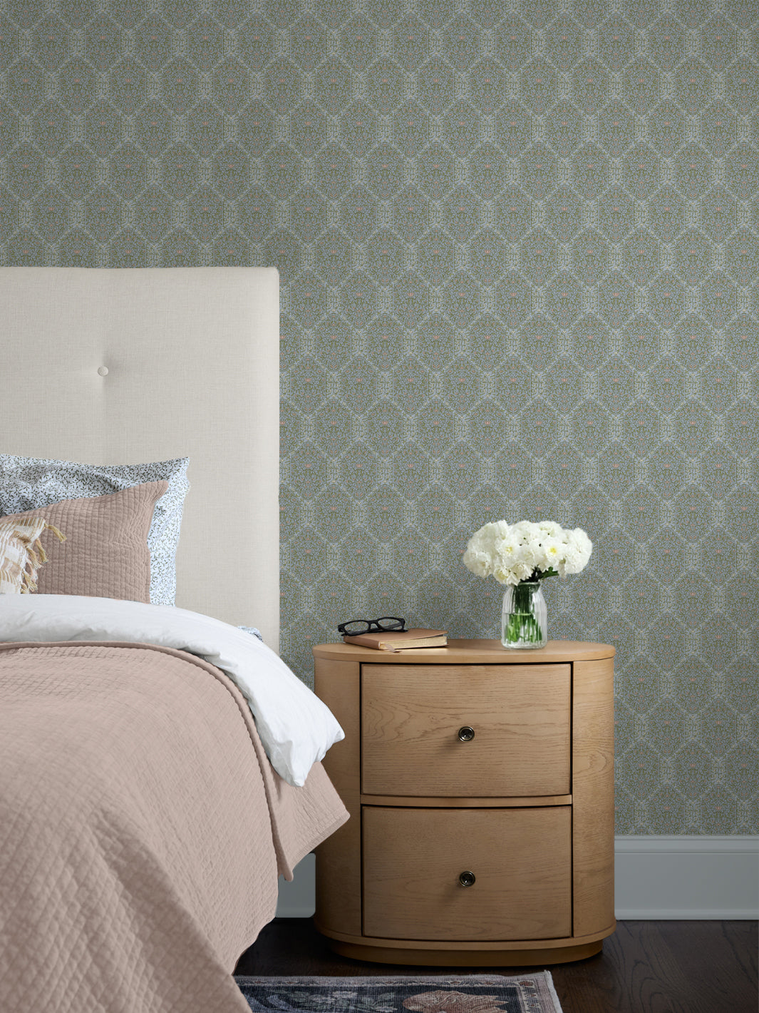 Blue-green Taliah wallpaper behind a cream headboard, blush quilt and oak nightstand